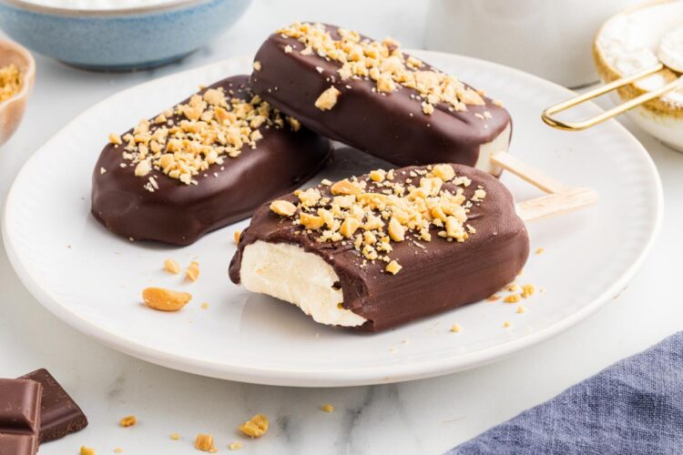 three chocolate-covered protein ice cream bars on a white plate with chocolate and peanuts in the background