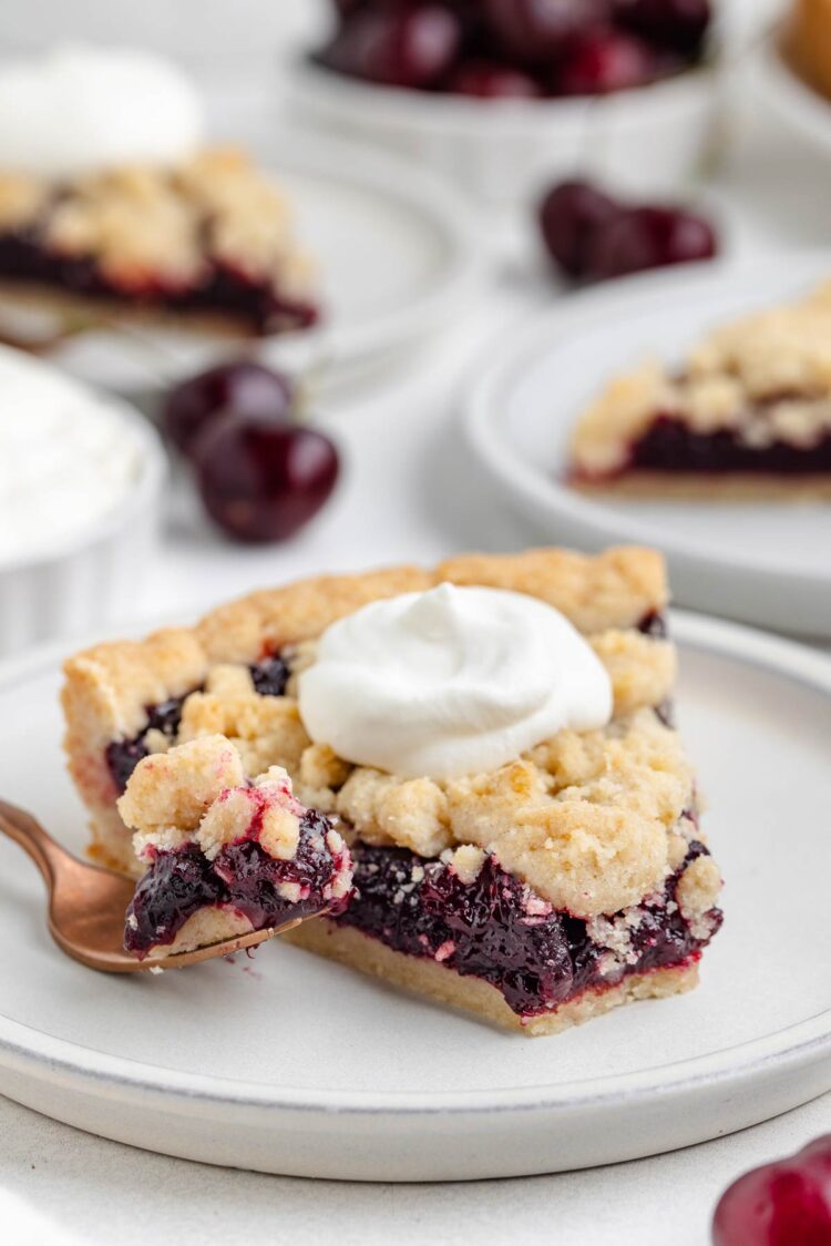 fork digging into a slice of cherry tart with whipped cream on top with more cherries on top in background