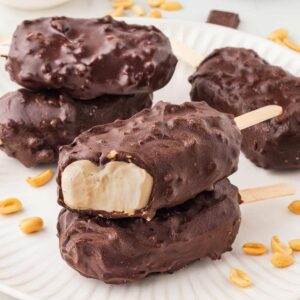 2 greek yogurt ice cream bars stacked on a white plate with more bars and peanuts in background