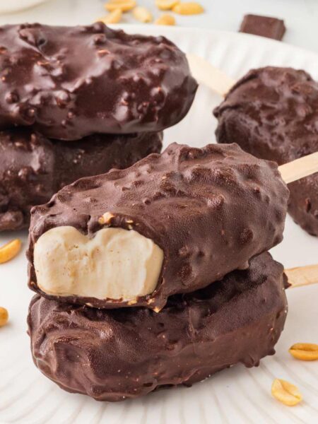 2 greek yogurt ice cream bars stacked on a white plate with more bars and peanuts in background