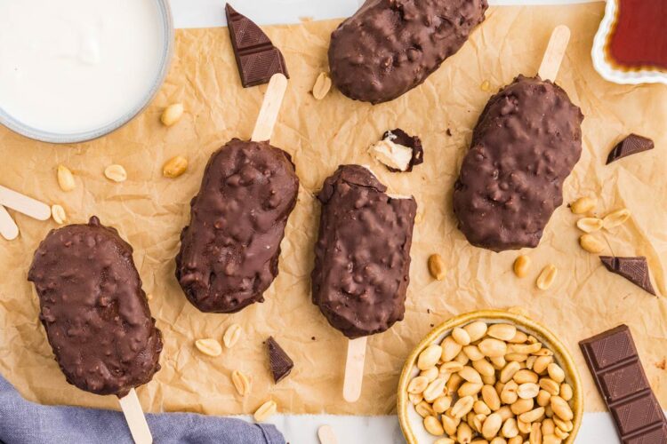 greek yogurt ice cream bars on a piece of parchment paper with a bowl of peanuts and melted chocolate in background