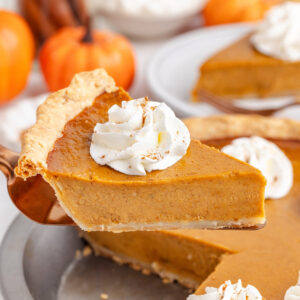 A slice of gluten-free pumpkin pie topped with whipped cream and cinnamon, served on a pie server with pumpkins in the background.