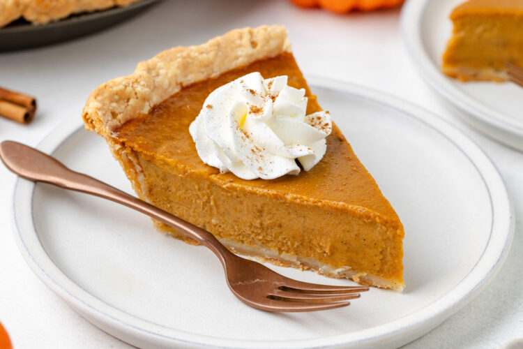 A slice of gluten-free pumpkin pie topped with whipped cream and cinnamon on a white plate with a copper fork.