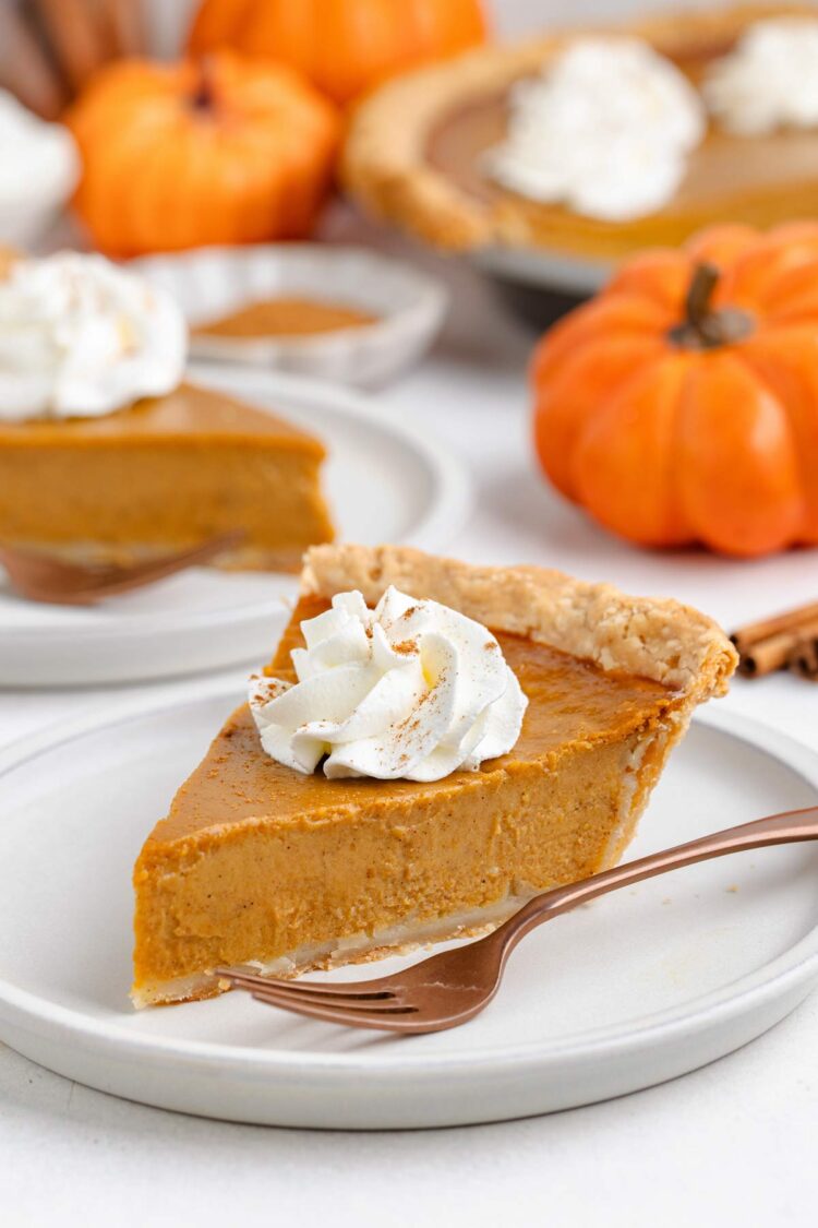 A slice of gluten-free pumpkin pie topped with whipped cream, surrounded by pumpkins and spices.