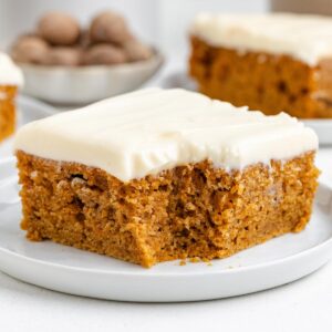 Moist sweet potato cake topped with creamy frosting, served on a white plate with a bowl of nuts in the background.