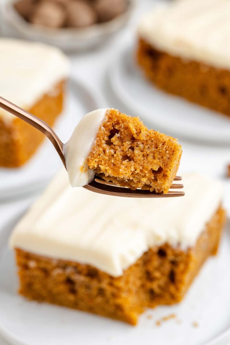 A fork holds a bite of sweet potato cake topped with cream cheese frosting, with cake slices in the background.