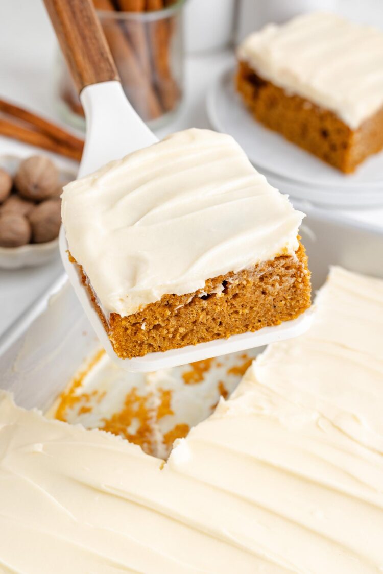 A slice of sweet potato cake with cream cheese frosting being lifted from a baking dish, surrounded by spices.